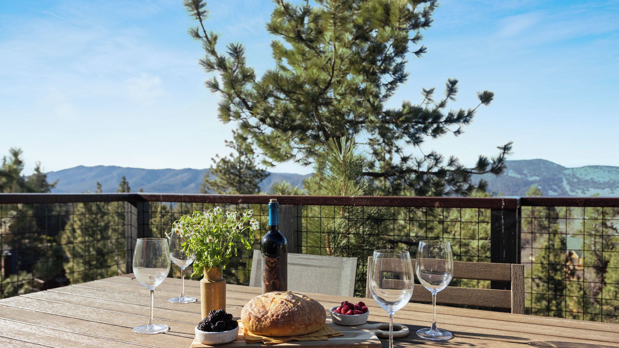 A wooden table with wine glasses, a loaf of bread, a bowl of berries, and a bottle of wine, set against a scenic mountain backdrop.