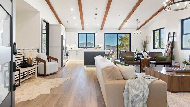 A spacious living area with a wooden ceiling, a large sofa, a coffee table, and a view of the kitchen and outdoors.