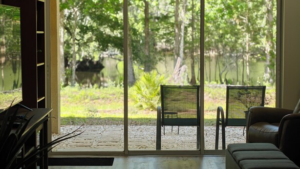 A room with a large sliding glass door leading to a garden with chairs and a view of trees and a pond. - Wise Owl Retreat, a beautiful suite amongst towering oaks, access to River/view. (Dunnellon)