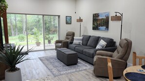 A modern living room with a grey sofa, a brown armchair, and a coffee table. There is a large window with a view of greenery outside. - Wise Owl Retreat, a beautiful suite amongst towering oaks, access to River/view. (Dunnellon)