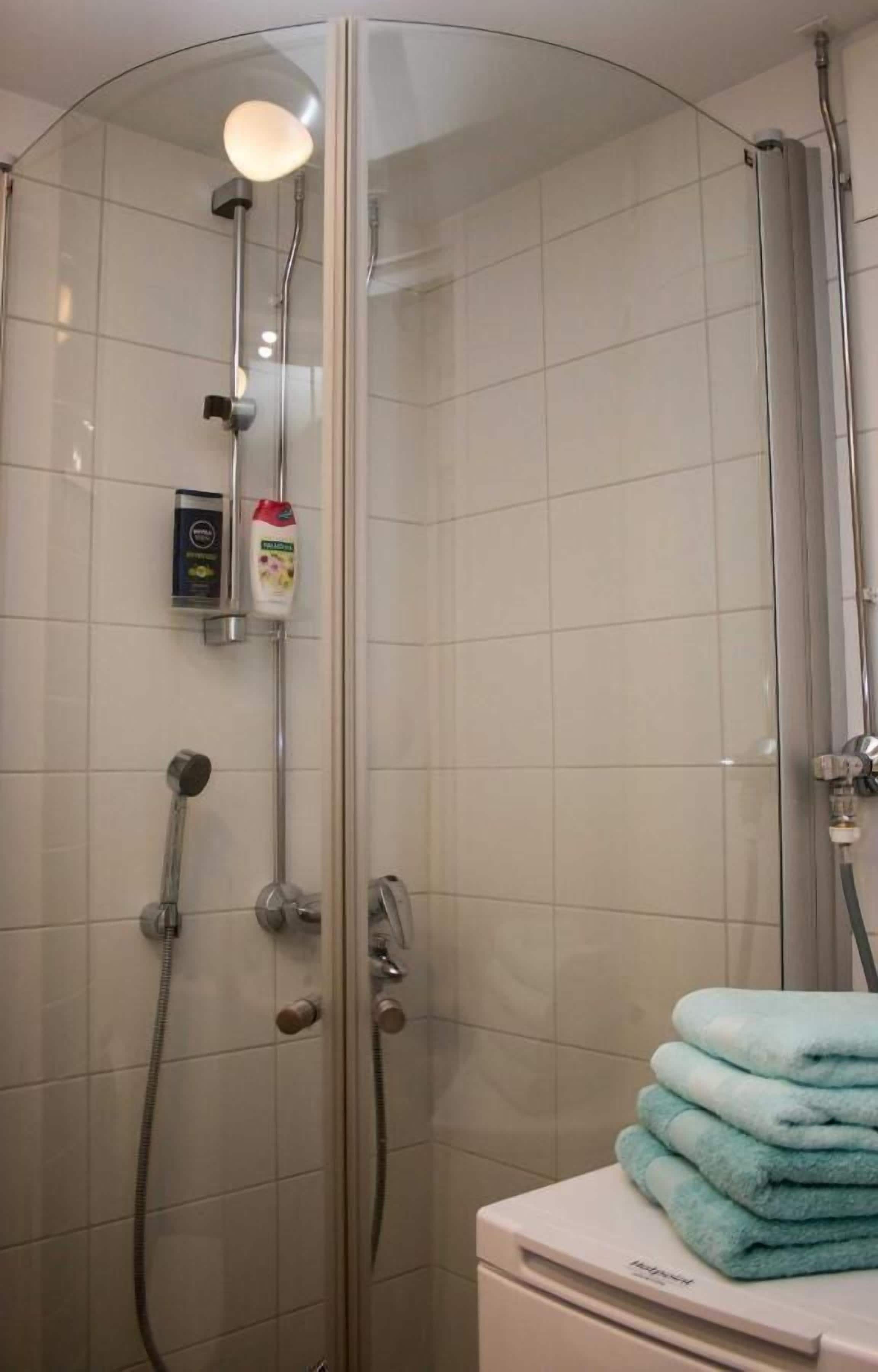 Une douche fermée par des parois en verre, dotée d’un pommeau de douche à main et d’un robinet mural. Un autre pommeau de douche à main est également présent. Une pile de serviettes est posée sur un comptoir blanc, à côté de la douche.