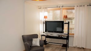 Un salon avec un fauteuil gris, un téléviseur  à écran plat posé sur un meuble noir, une petite table avec une lampe et une fenêtre équipée de stores.