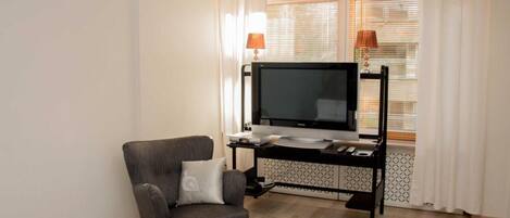 A living room with a grey armchair, a flat-screen TV on a black stand, a small table with a lamp, and a window with blinds.
