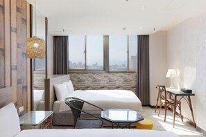 A modern hotel room with a large bed, a seating area with a glass table, a small side table, and a view of the cityscape through the window.