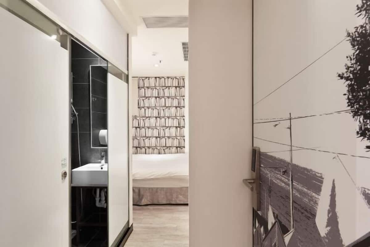 Un couloir étroit mène à une chambre à coucher avec un lit, une salle de bain avec un lavabo et un mur orné d’un motif noir et blanc.