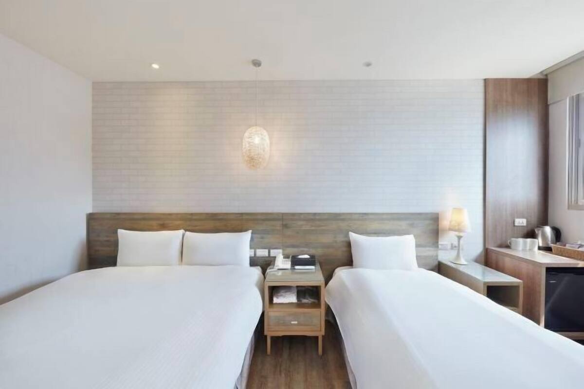 Une chambre d’hôtel avec deux lits, une tête de lit en bois, une table de chevet avec une lampe et une petite kitchenette.