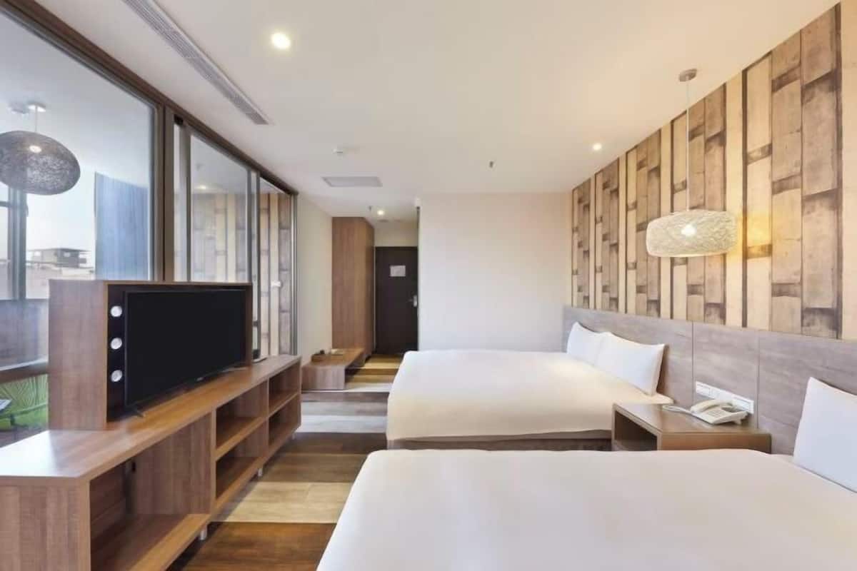 Une chambre d’hôtel avec deux lits, un téléviseur posé sur une console en bois, une lampe fixée au mur et une vue dégagée sur l’extérieur grâce à de grandes fenêtres.