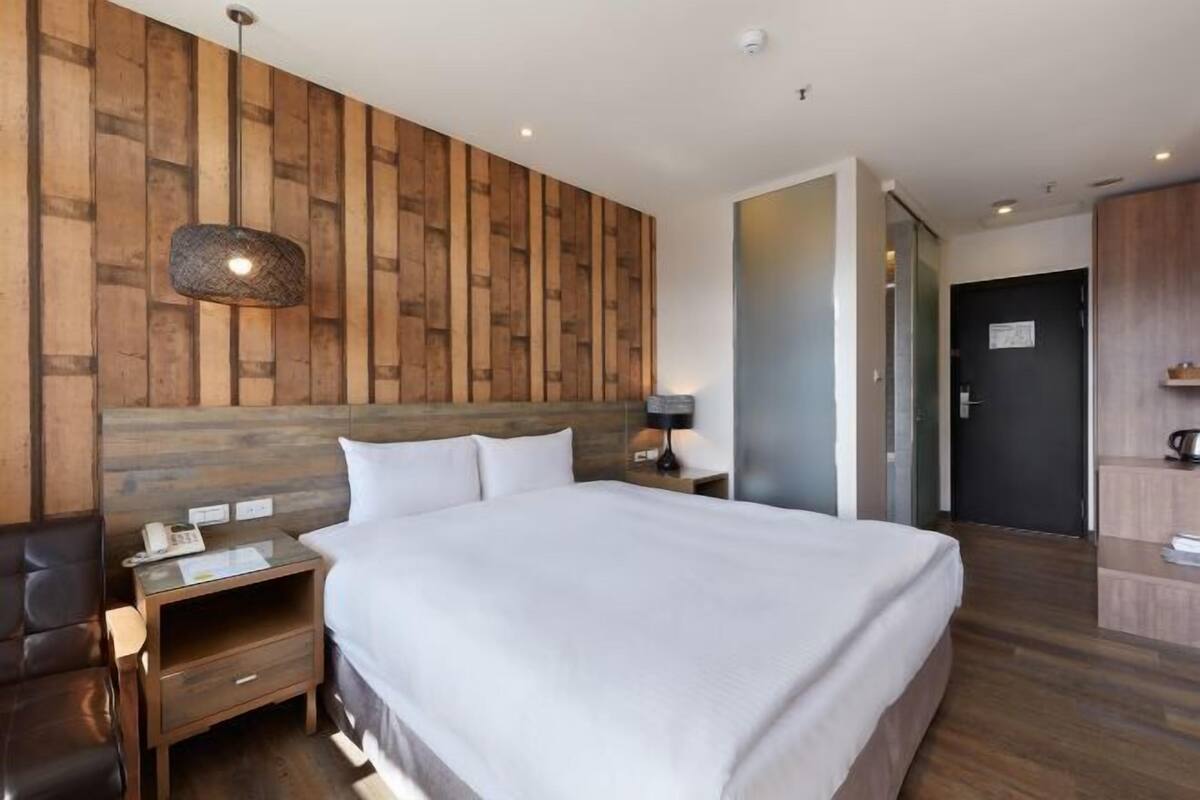 Une chambre d’hôtel moderne avec une tête de lit en bois, un grand lit, une table de chevet avec une lampe et un petit bureau avec un téléphone.