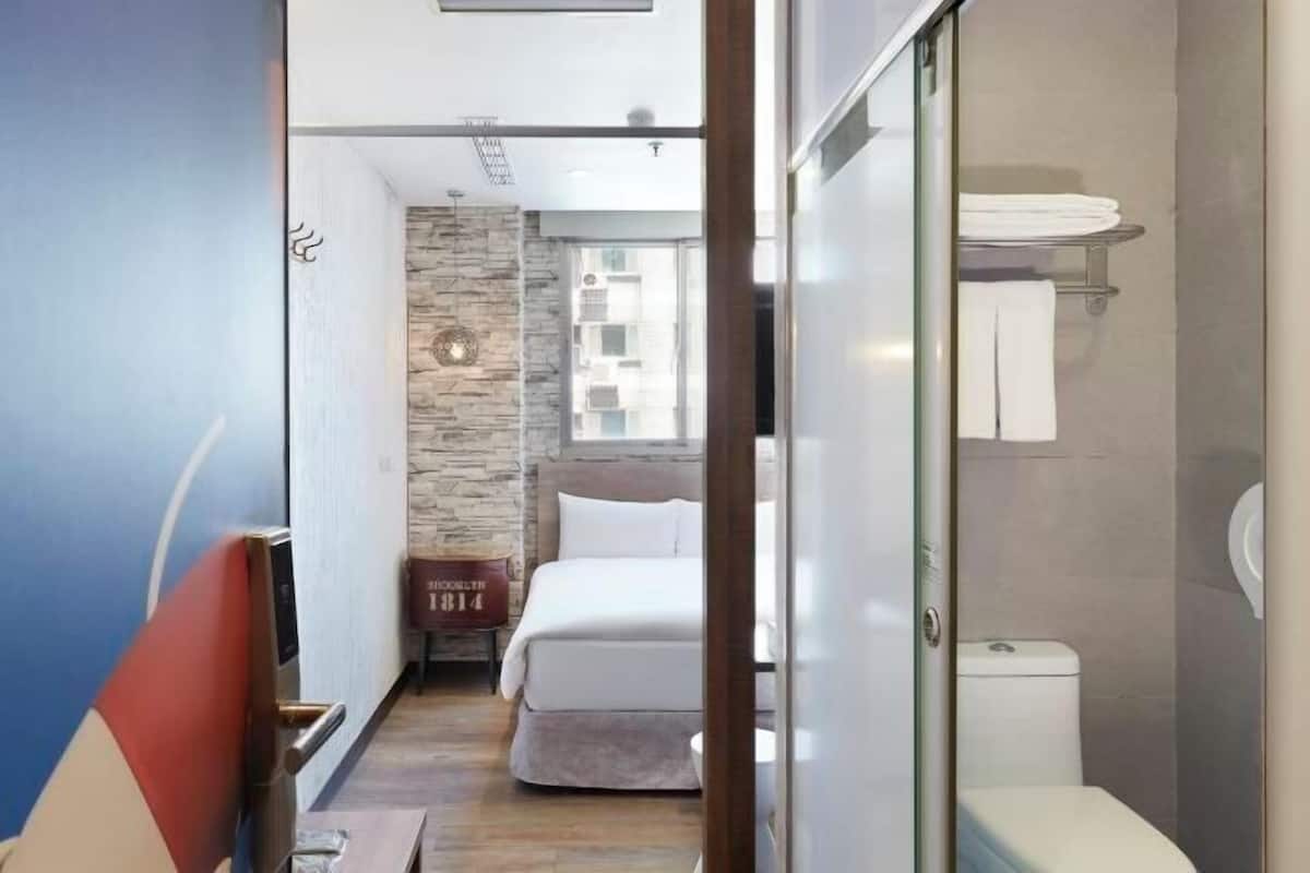 Une chambre d’hôtel étroite, avec un lit, une petite table, une cabine de douche en verre et une vue sur l’extérieur.