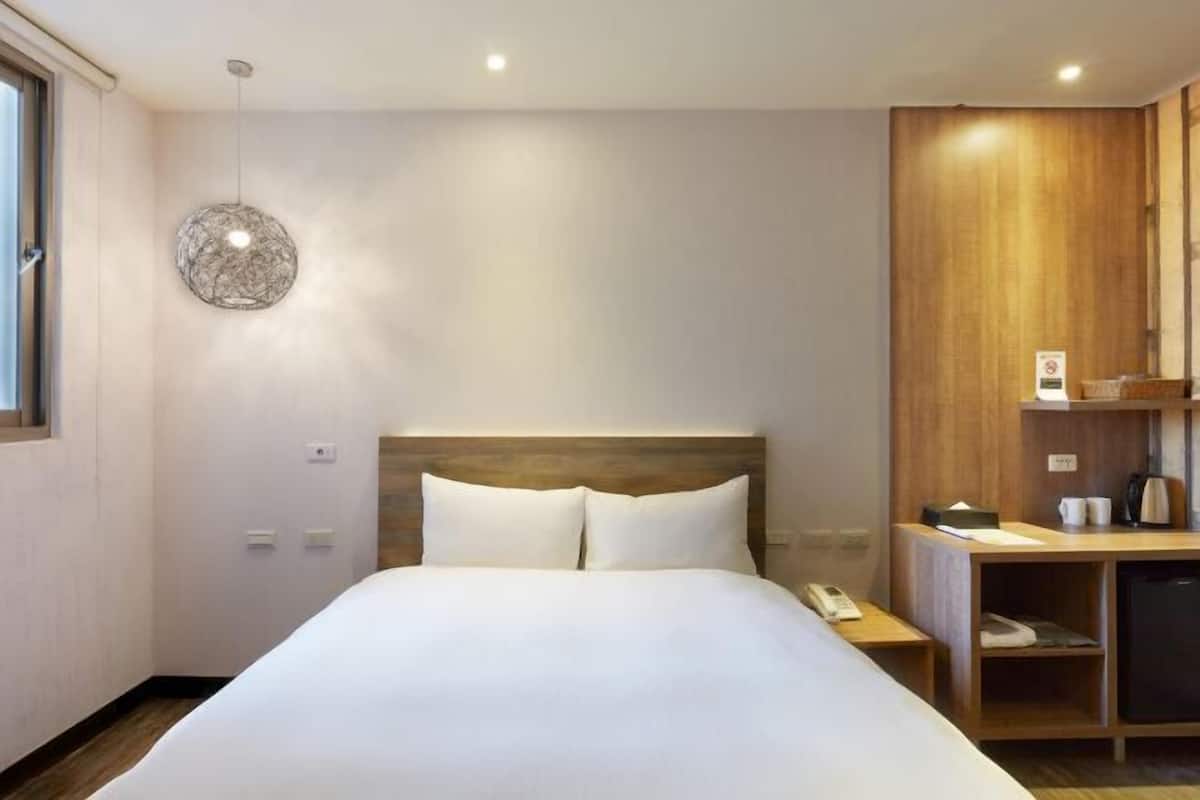 Une chambre d’hôtel avec un grand lit, une tête de lit en bois, une table de chevet avec un téléphone, une petite armoire et un luminaire fixé au mur.