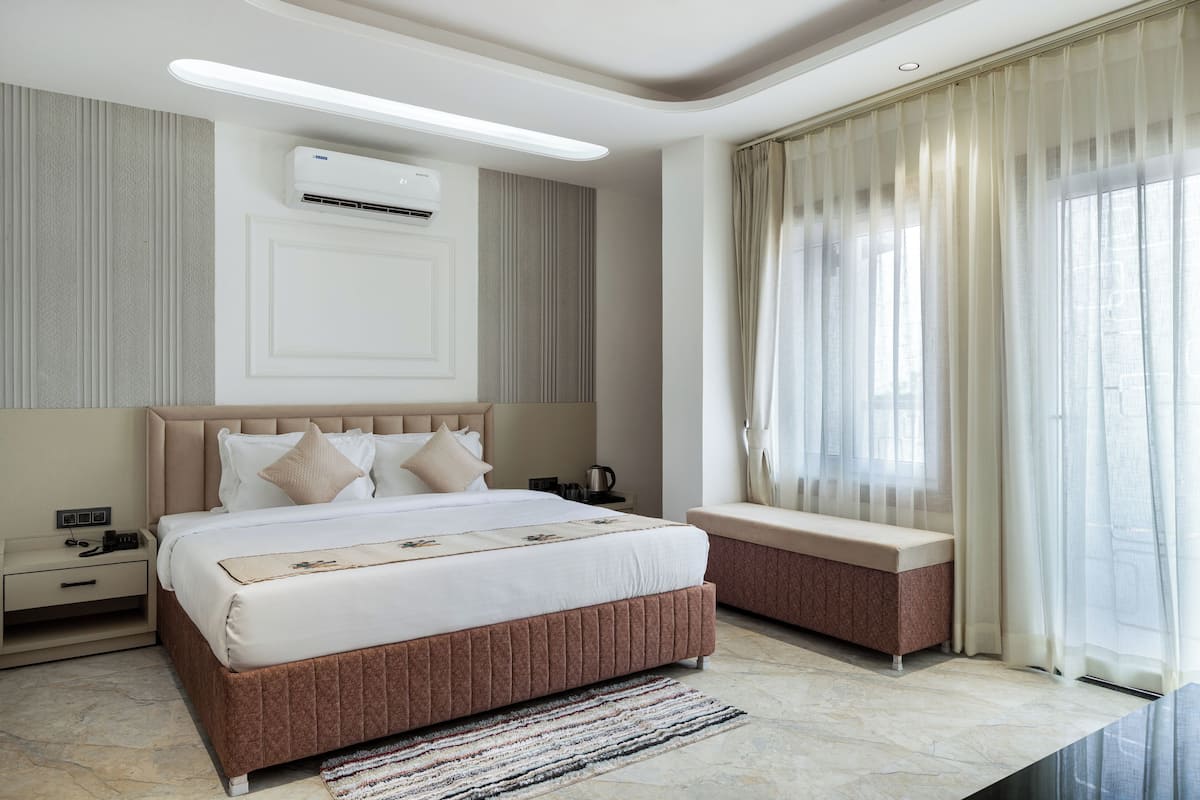 A modern hotel bedroom with a large bed, bedside tables, an air conditioning unit, and a view of the city through sheer curtains.