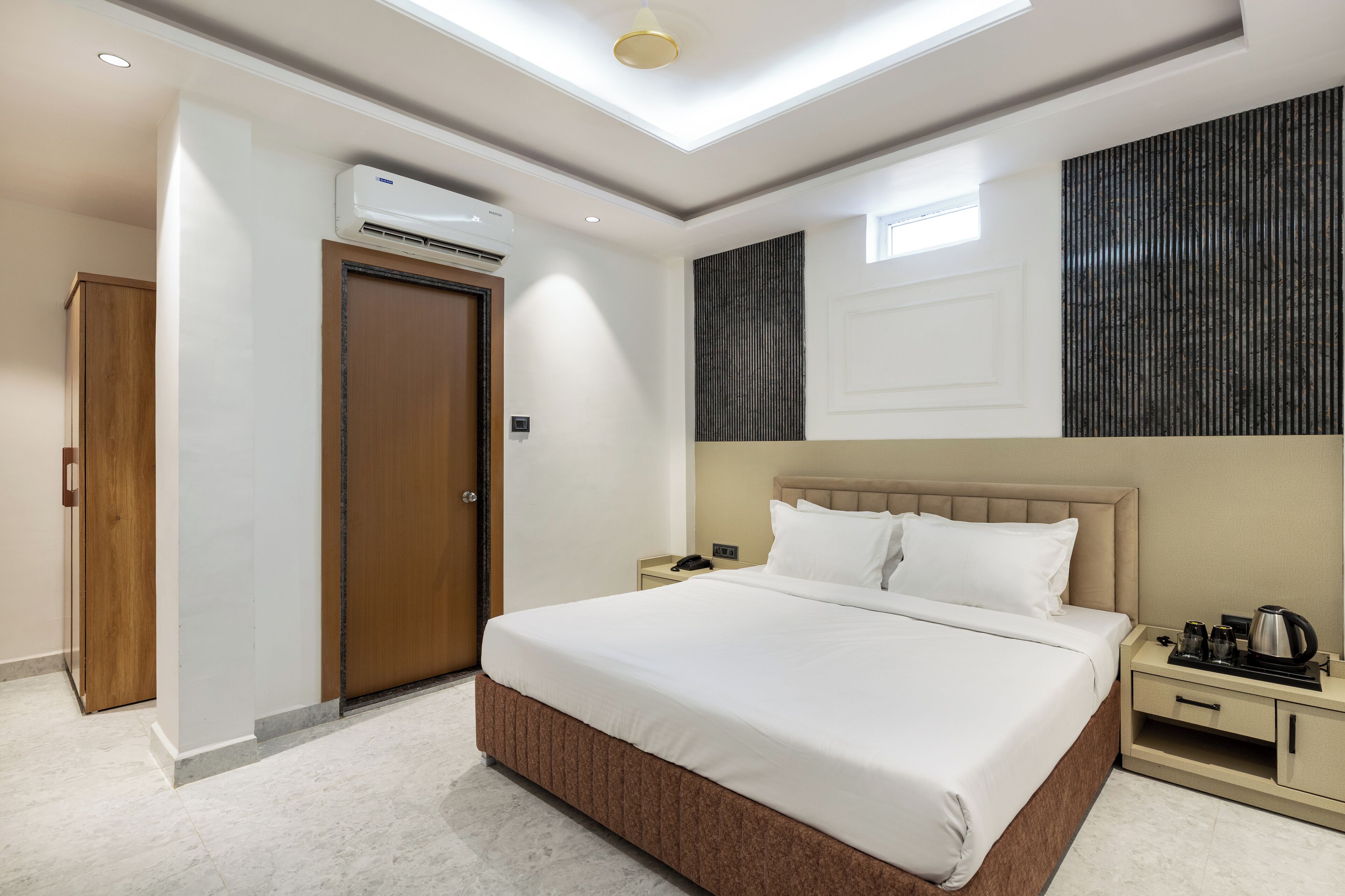 A hotel room with a large bed, a bedside table with a phone and a kettle, a wardrobe, and an air conditioning unit.