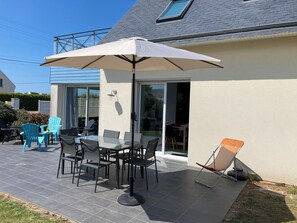 Um pátio externo com mesa, cadeiras, um guarda-sol grande e uma porta de vidro deslizante que dá para um espaço interno.