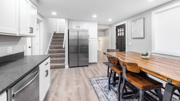 A modern kitchen with white cabinets, a stainless steel refrigerator, a wooden dining table, and a staircase leading to another level. - Newly Renovated 4 bedroom home (West Valley City)