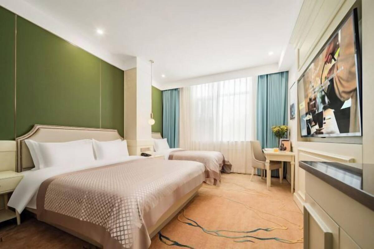 A hotel room with two beds, a desk, a chair, a TV, and a large window with curtains.