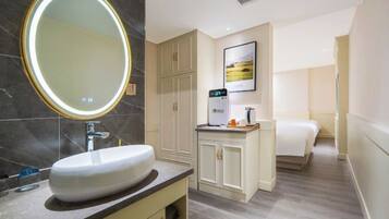 A modern bathroom with a large oval mirror, a white sink, and a built-in cabinet. There is a bed with a white duvet and a small bedside table with a lamp.