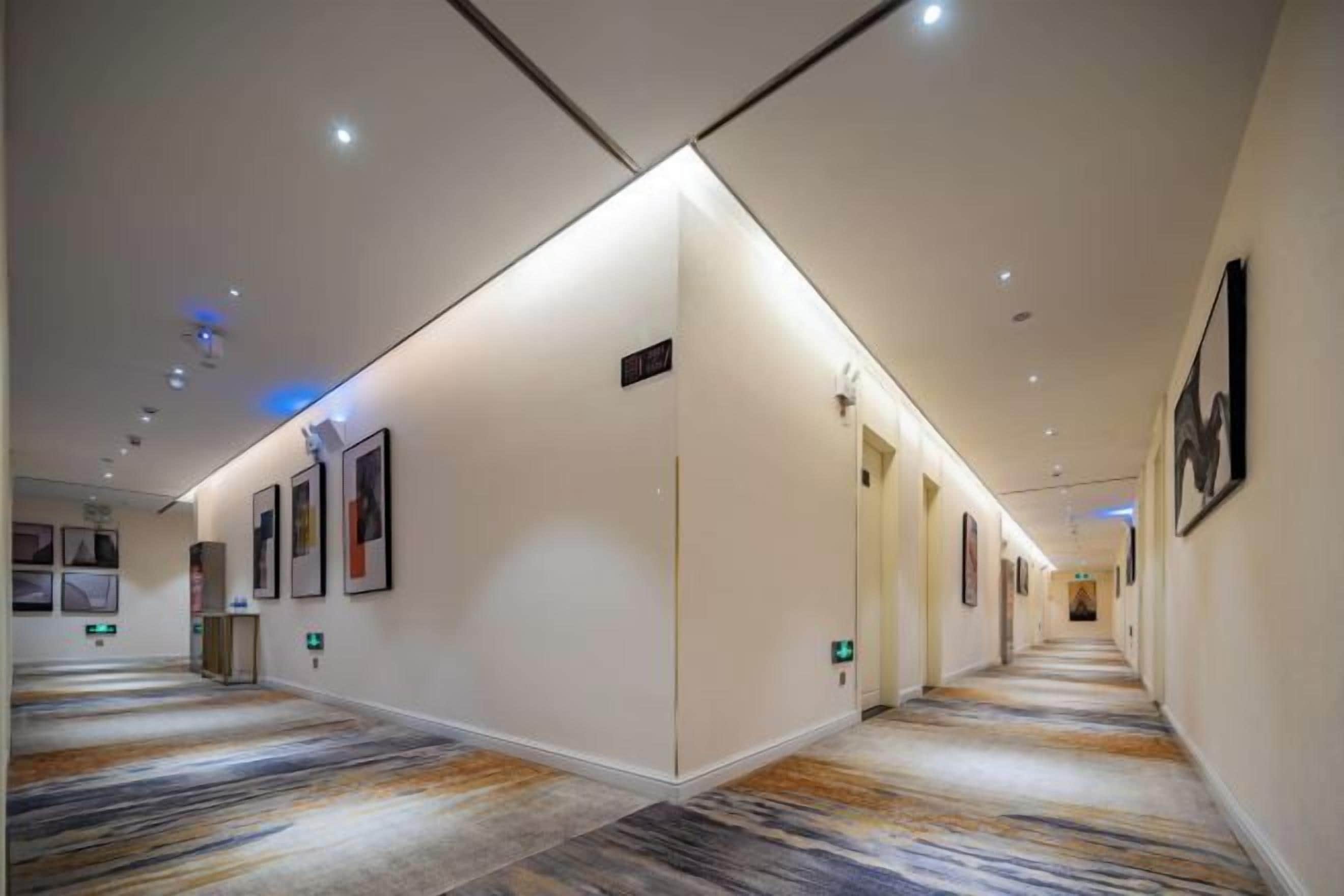 A hotel corridor with a carpeted floor, framed artwork on the walls, and a well-lit ceiling.
