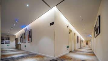 A hotel corridor with a carpeted floor, framed artwork on the walls, and a well-lit ceiling.