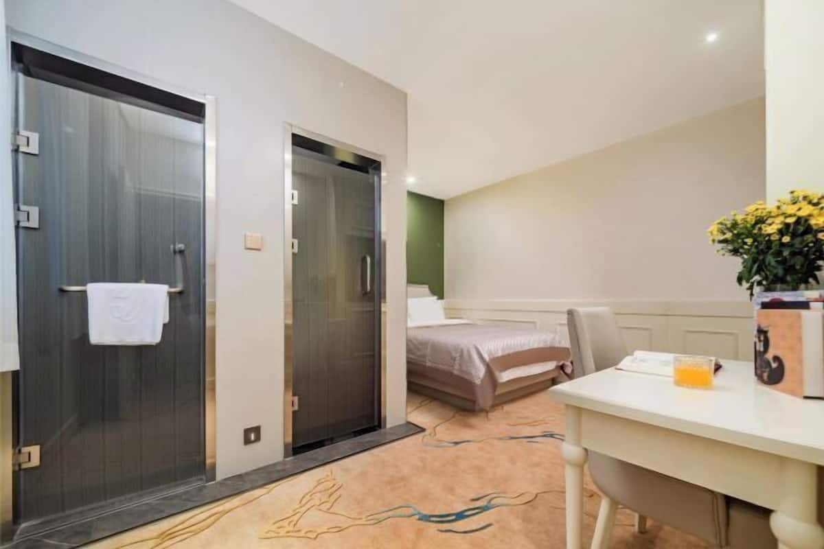 A hotel room with a bed, a desk with a chair, a shower, and a small table with a vase of flowers.