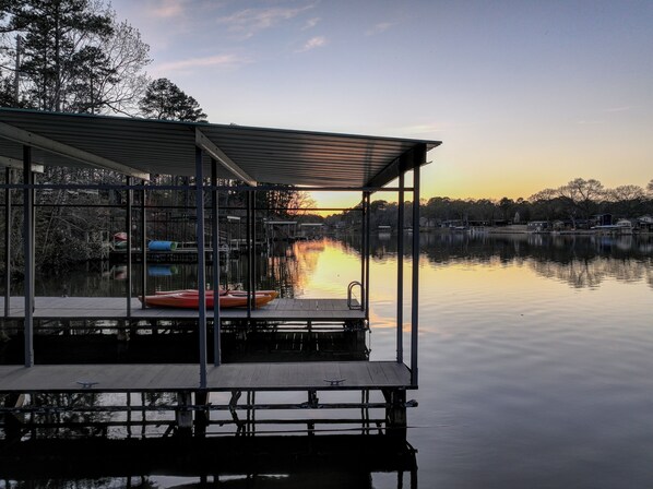 Un muelle con una zona de estar cubierta, un lago tranquilo y una puesta de sol al fondo.