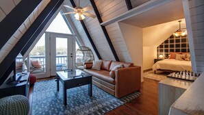 A cozy attic bedroom with a loft bed, a leather sofa, a chessboard, and a large window overlooking a balcony.
