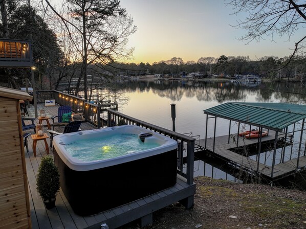 A hot tub on a wooden deck overlooking a calm lake with docked boats, surrounded by trees and a wooden cabin.