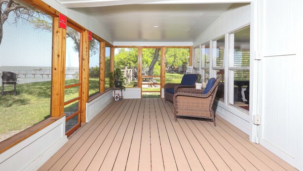 A covered outdoor deck with wooden flooring, wicker furniture, and large windows offering a view of the outdoors.