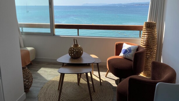 A room with a view of the sea, a round table, a brown armchair, and a woven floor lamp. - Waterfront studio with enchanting views (Six-Fours-les-Plages)