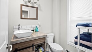 Een badkamer met een houten wastafelmeubel, een witte wastafel, een toilet en een raam met luxaflex.
