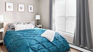 Een slaapkamer met een groot bed, een nachtkastje met een lamp, een raam met luxaflex en drie ingelijste foto's aan de muur.