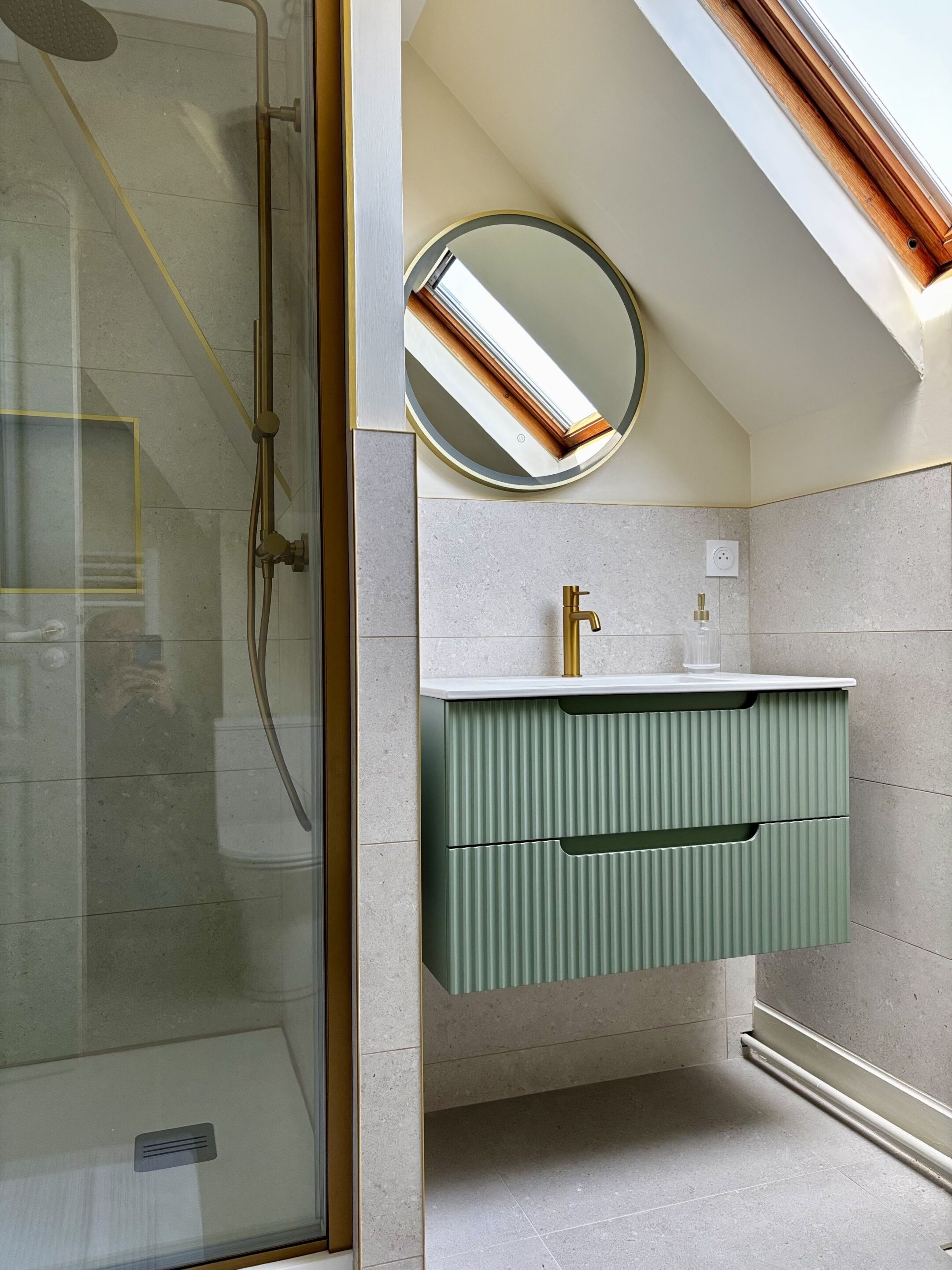 A bathroom with a glass shower enclosure, a round mirror, a green vanity with a gold faucet, and a white countertop.