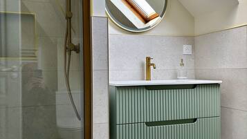 A bathroom with a glass shower enclosure, a round mirror, a green vanity with a gold faucet, and a white countertop.