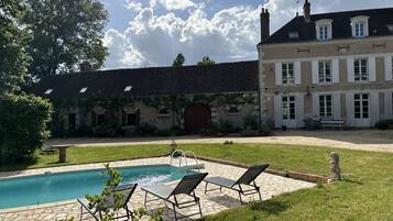 A swimming pool with lounge chairs, a stone patio, and a large building with a tiled roof and multiple windows.