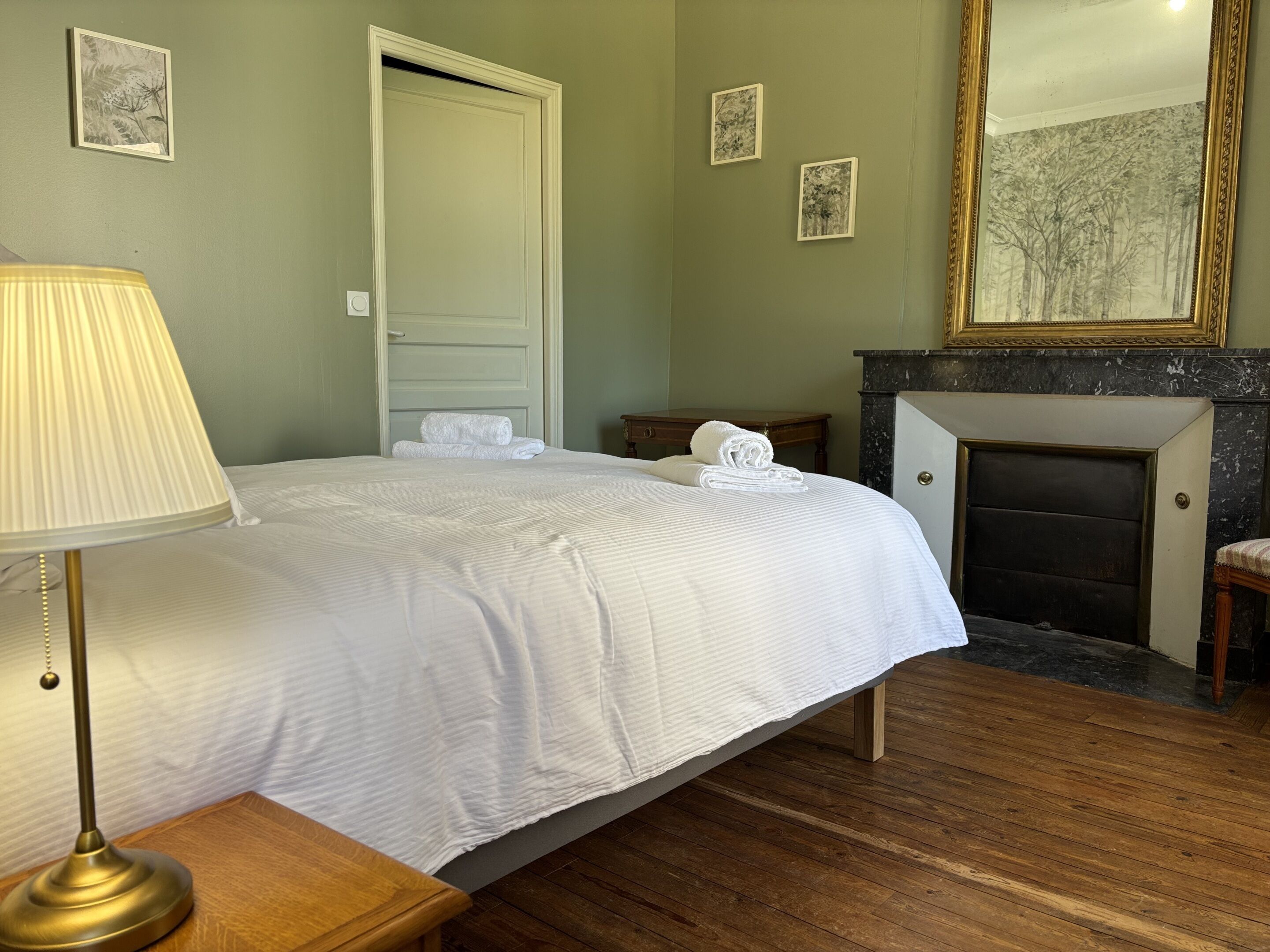 Una cama bien hecha con sábanas blancas, una lámpara de noche, un mesita de madera, una pequeña mesa con lámpara y una chimenea con detalles de mármol.