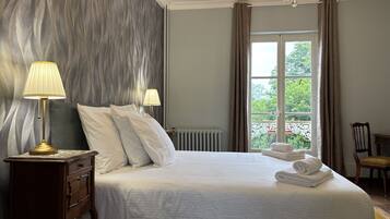 A neatly made bed with white linens, two bedside lamps, a wooden nightstand, and a view of greenery through a window.