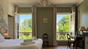 Ένα υπνοδωμάτιο με ένα μεγάλο κρεβάτι, δύο παράθυρα με κουρτίνες, μια ξύλινη καρέκλα και ένα γραφείο με ένα φωτιστικό.