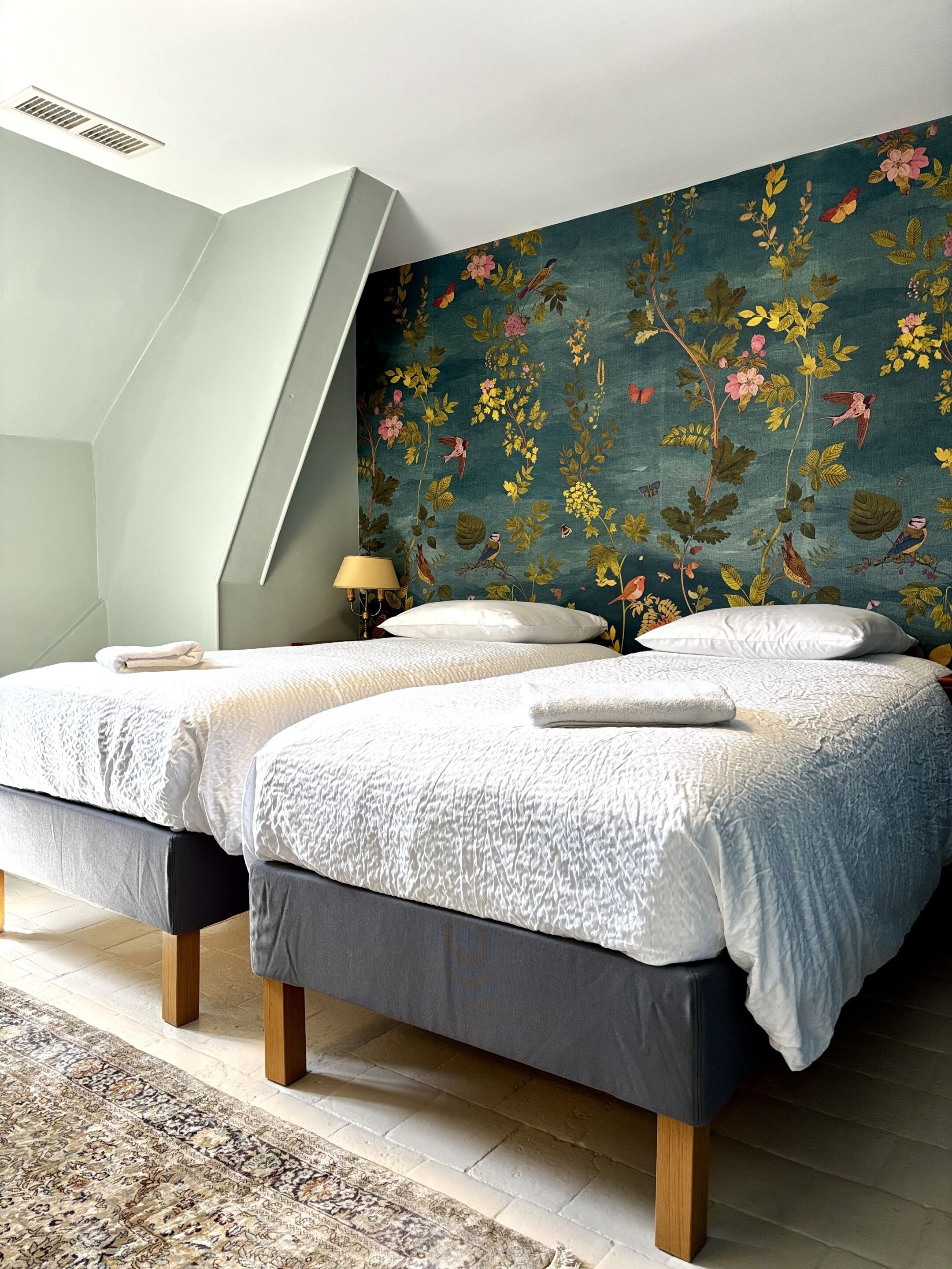 A bedroom with two beds, a patterned wall, a lamp, and a patterned rug.