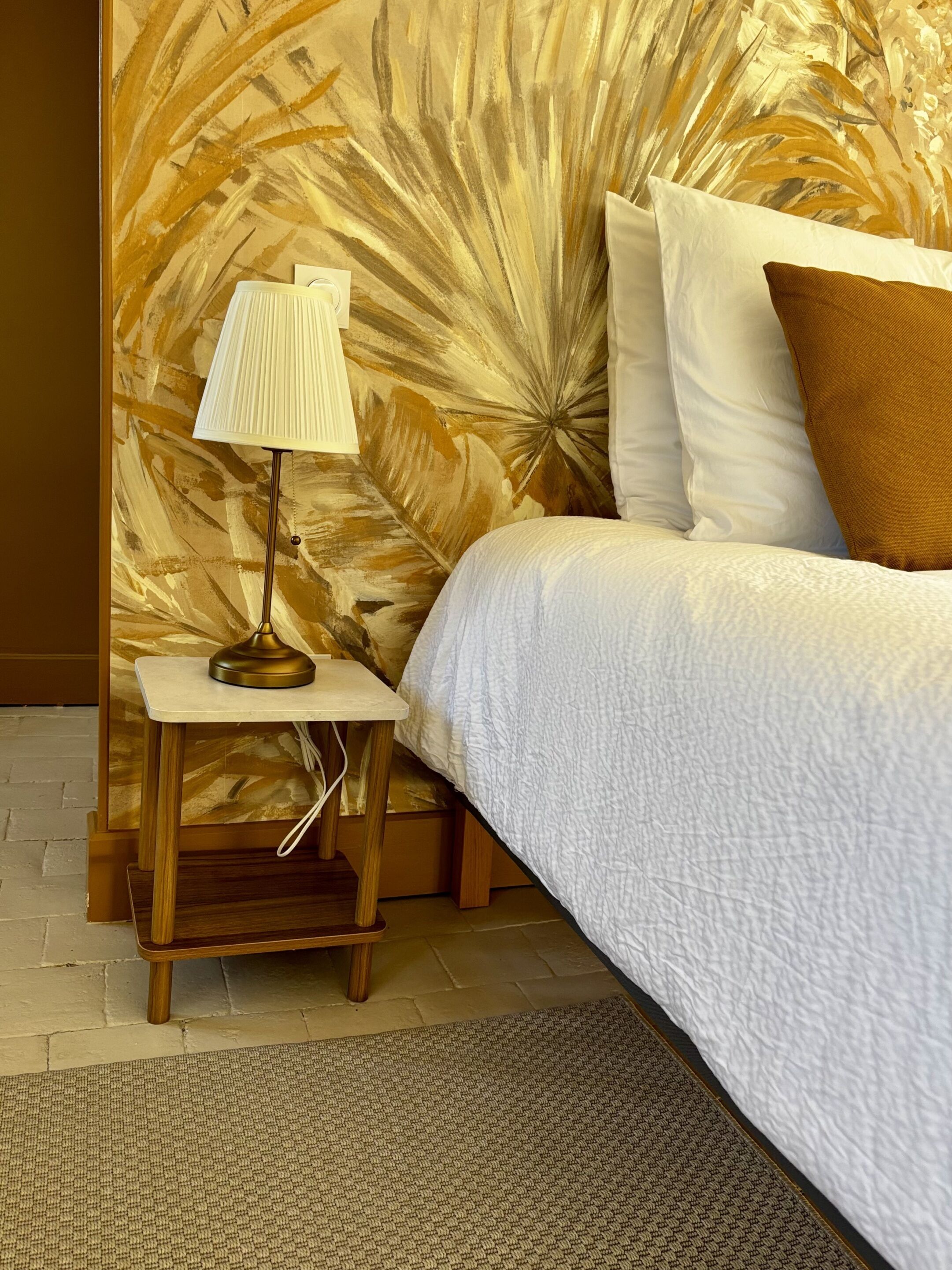 Una cama con colcha blanca, una lámpara de noche y una pared con diseño de hojas.