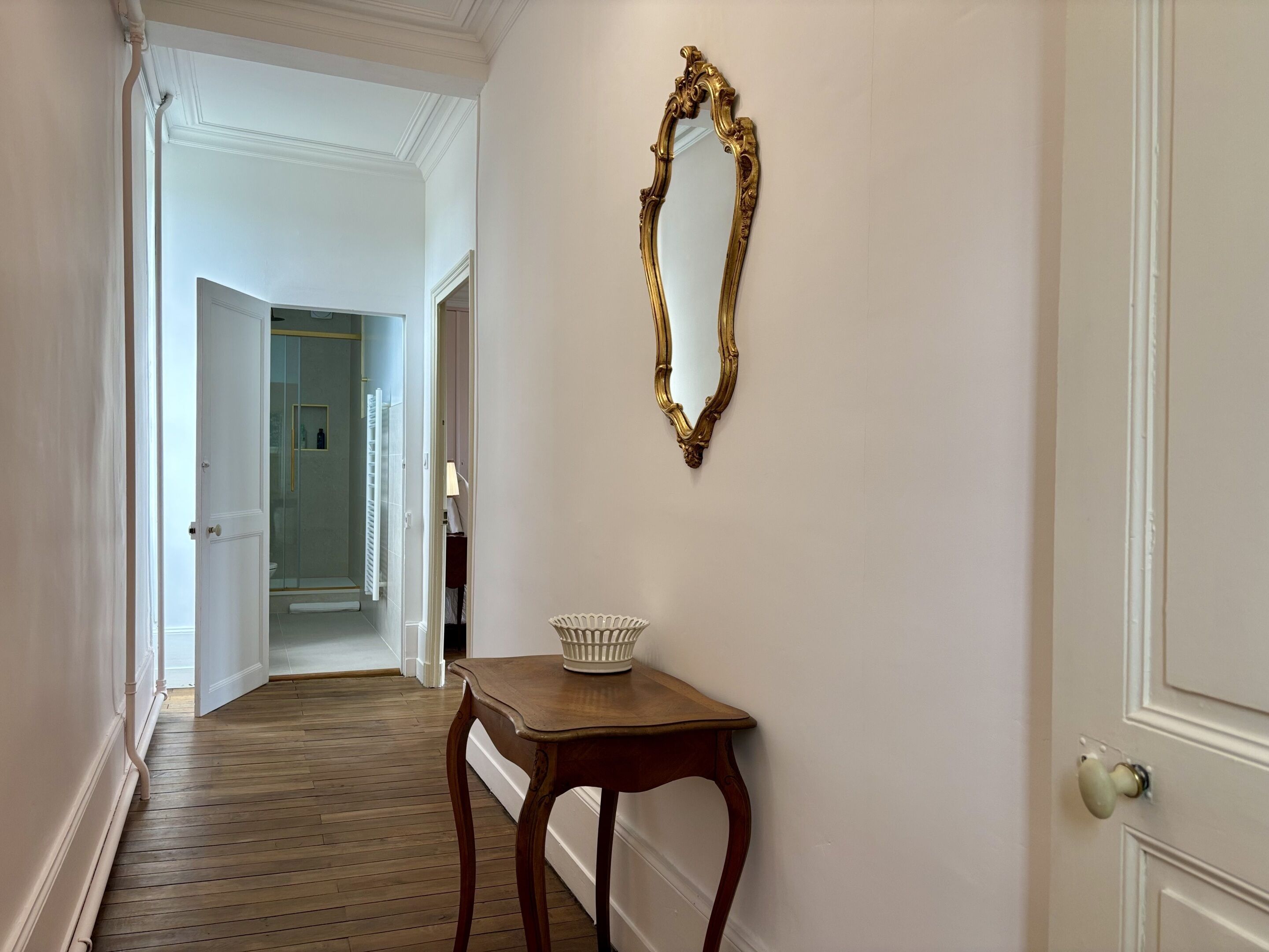 A hallway with a wooden floor, a white door, and a gold-framed mirror on the wall.