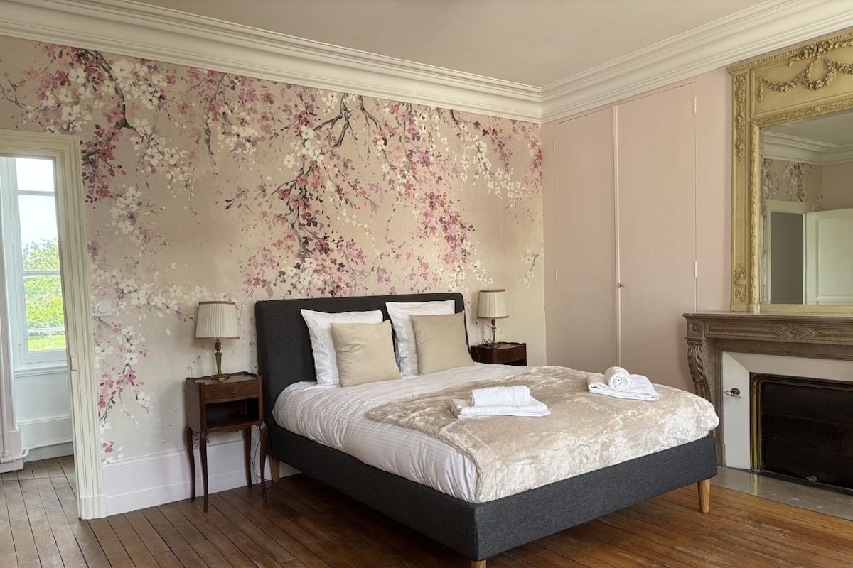 Un dormitorio con una cama grande, dos mesitas de noche, una chimenea y un papel tapiz con motivos florales.