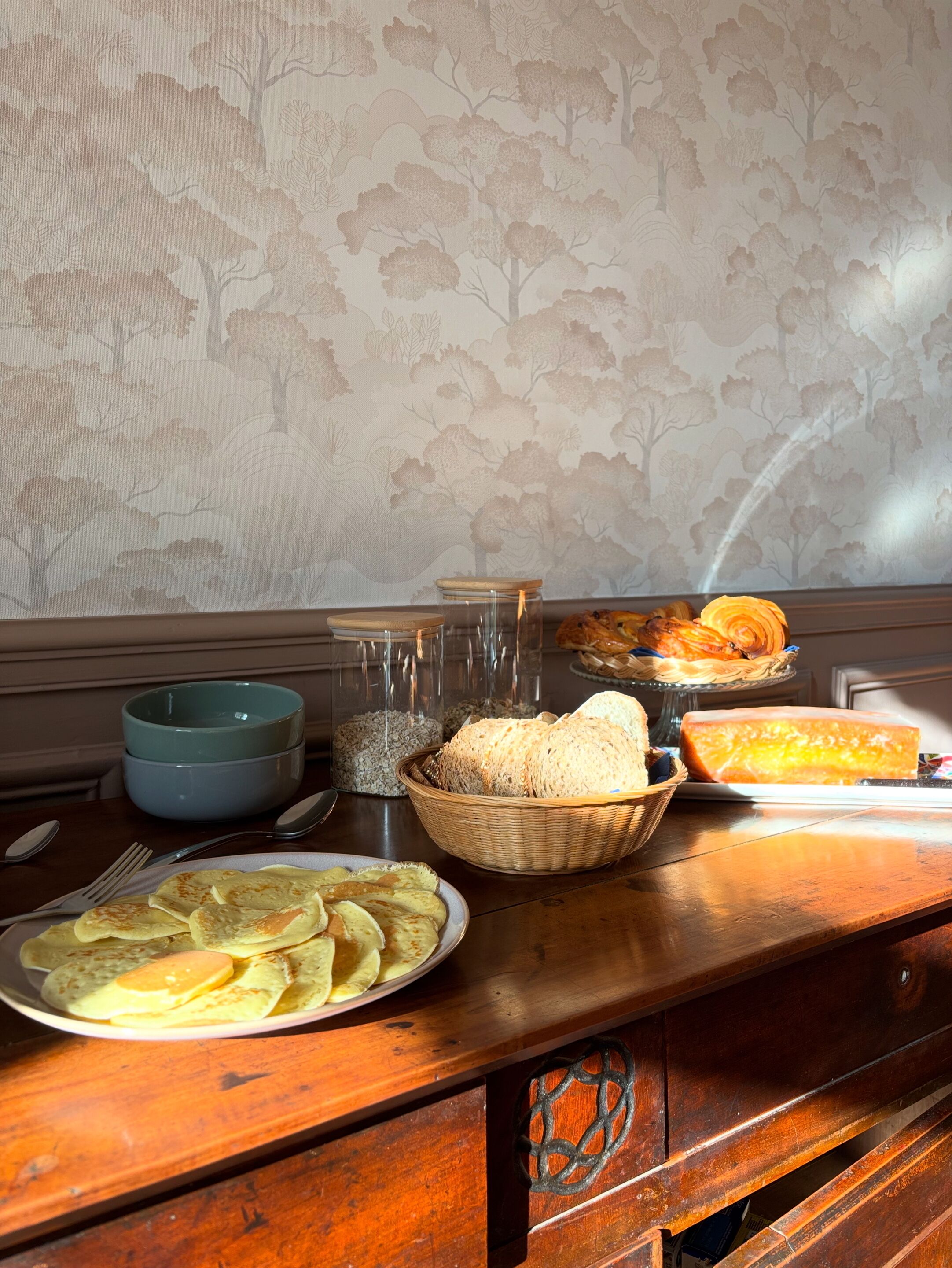 Une table à manger en bois, avec un papier peint à motifs floraux en arrière-plan, un bol bleu, un panier de pain et une assiette de crêpes.