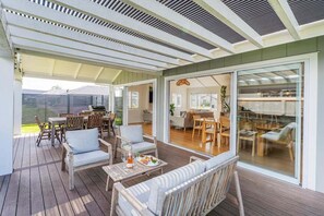 Ein möblierter Patio mit Holzmöbeln, einem Esstisch und einer Glastür zu einem Innenbereich mit Essplatz.