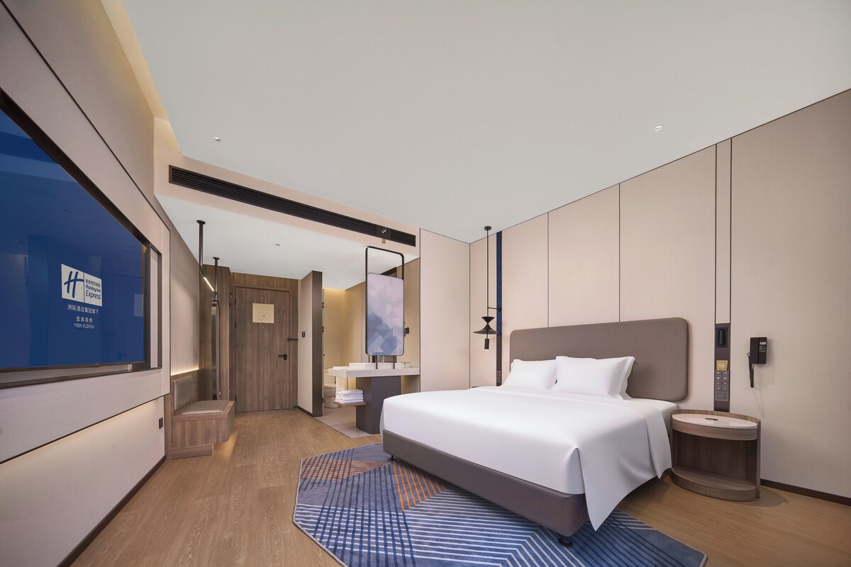 Una habitación de hotel moderna con una cama grande, una mesita de noche, un escritorio integrado, un espejo y una ventana con vistas al exterior.