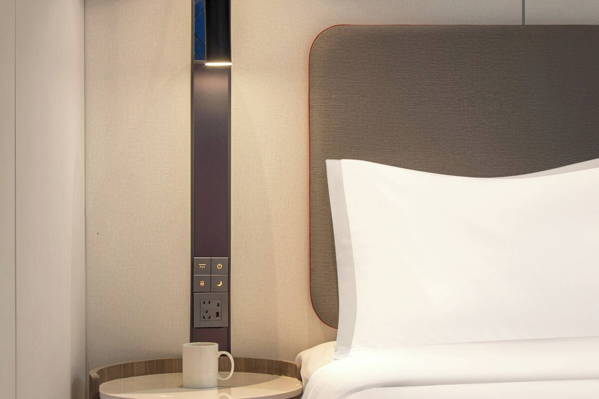 A hotel room with a bed, a bedside table with a cup, and a control panel with various buttons.