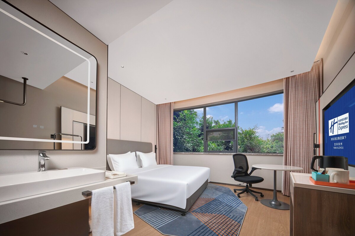 A hotel room with a bed, a desk, a chair, a large window, and a bathroom with a mirror and sink.