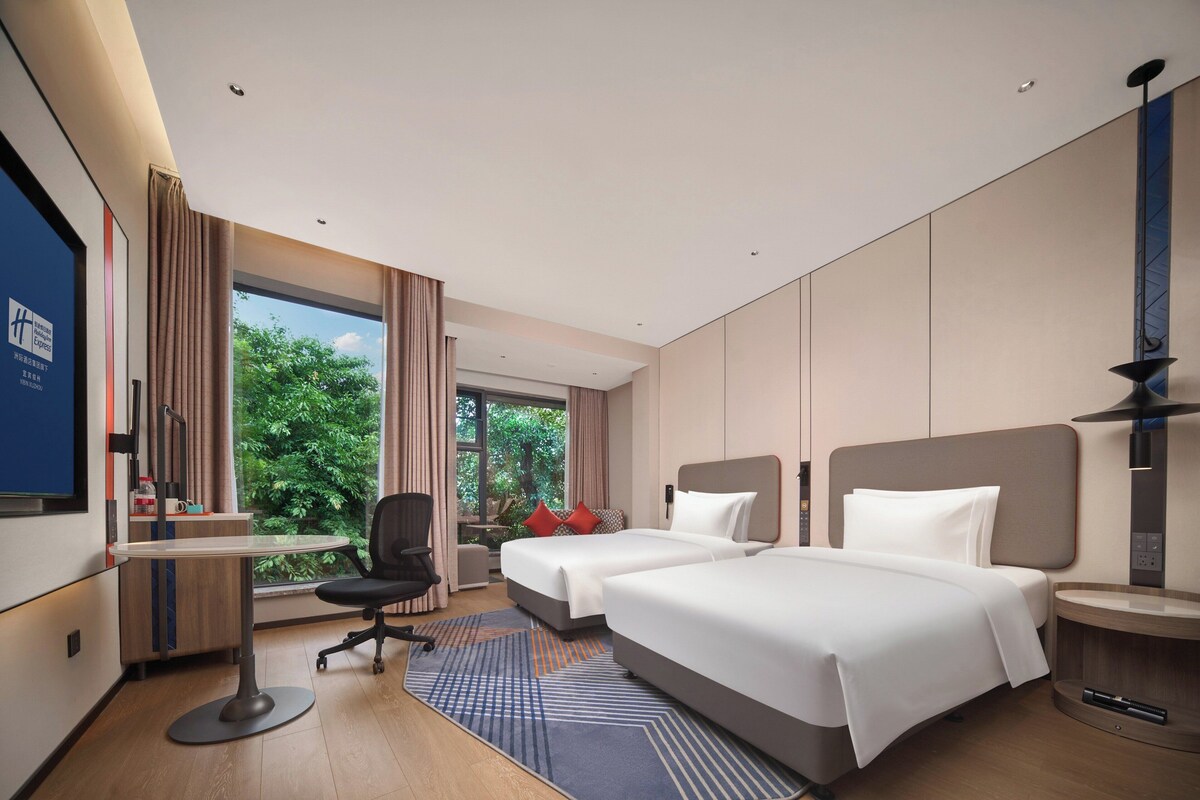 Une chambre d’hôtel avec deux lits, un bureau, une chaise et une grande fenêtre donnant sur un paysage verdoyant.