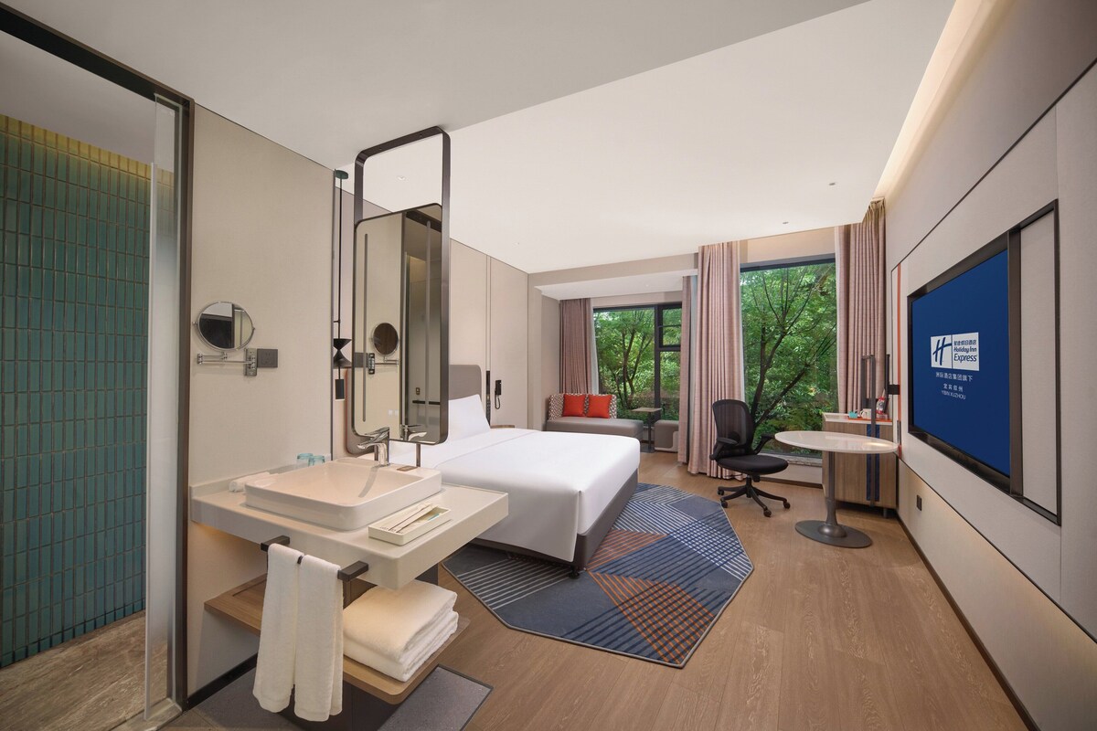 Une chambre d’hôtel moderne avec un grand lit, une salle de bain équipée d’un lavabo et d’un miroir, un bureau avec une chaise et un téléviseur fixé au mur.