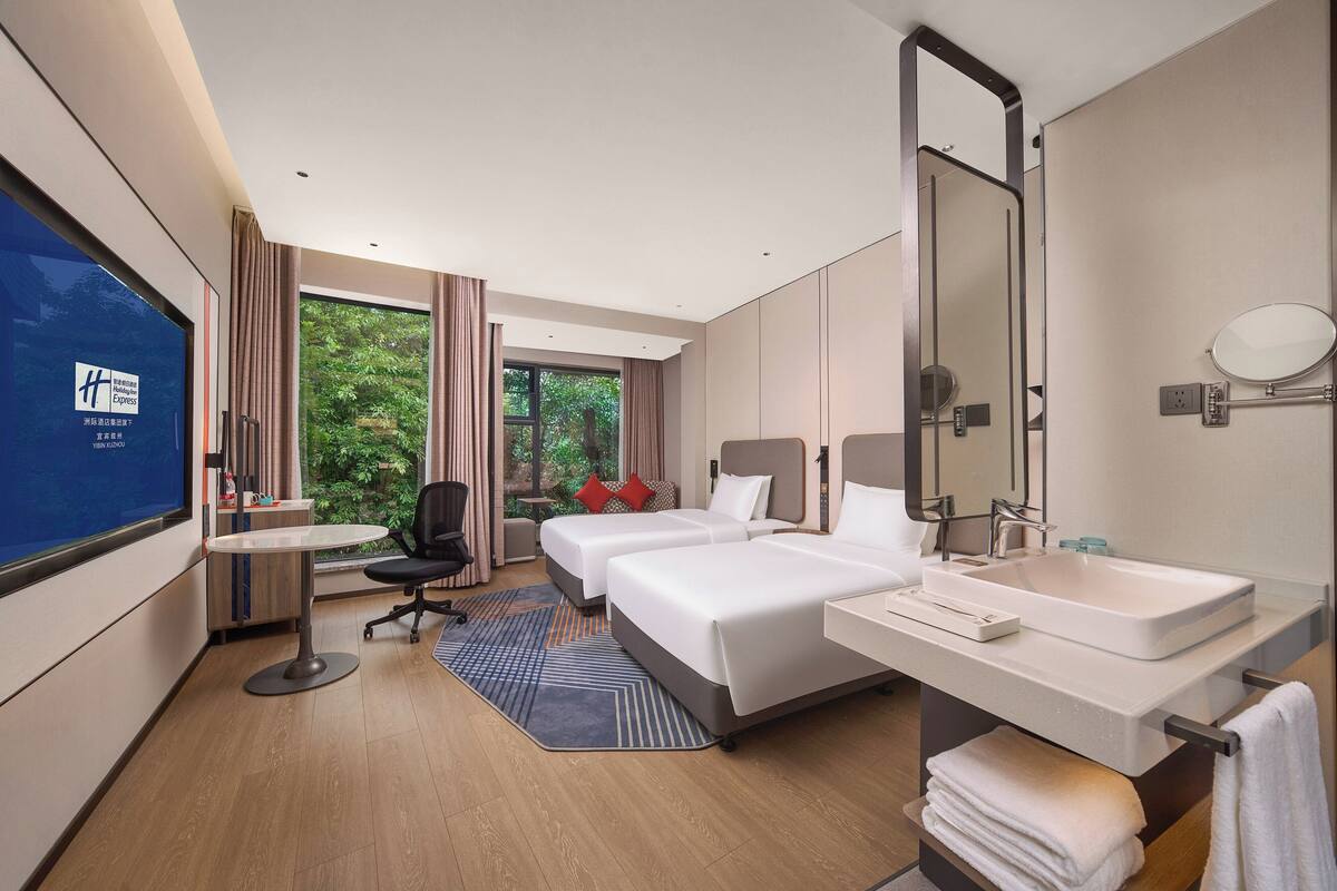 A hotel room with two beds, a desk, a large window with curtains, a bathroom with a sink and mirror, and a TV on the wall.