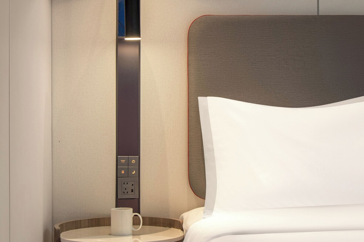 Una habitación de hotel con una cama, una mesita de noche con una taza y un panel de control con varios botones.