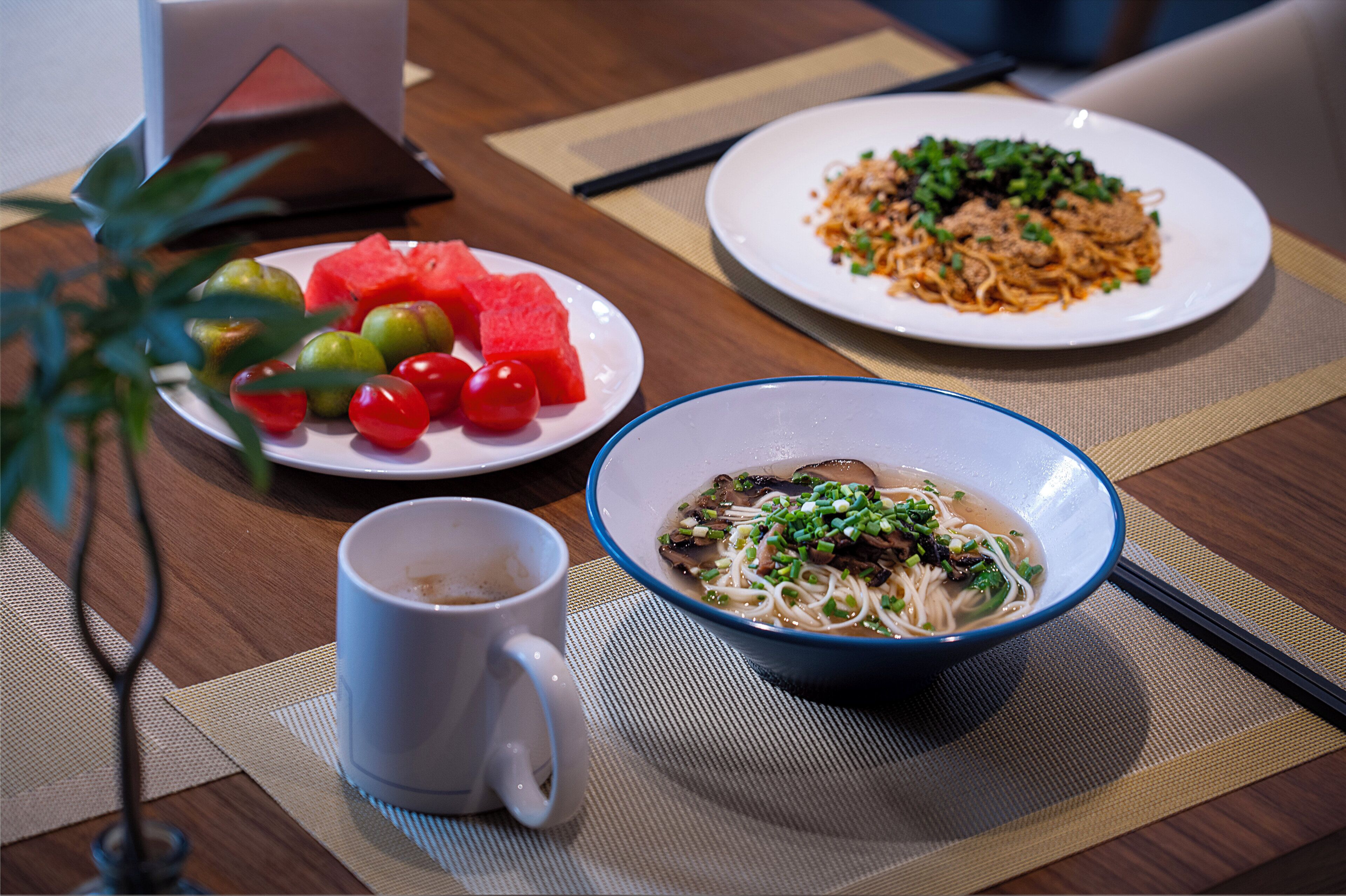 A table with a bowl of noodle soup, a plate of watermelon, cherry tomatoes, and a plate of stir-fried noodles.
